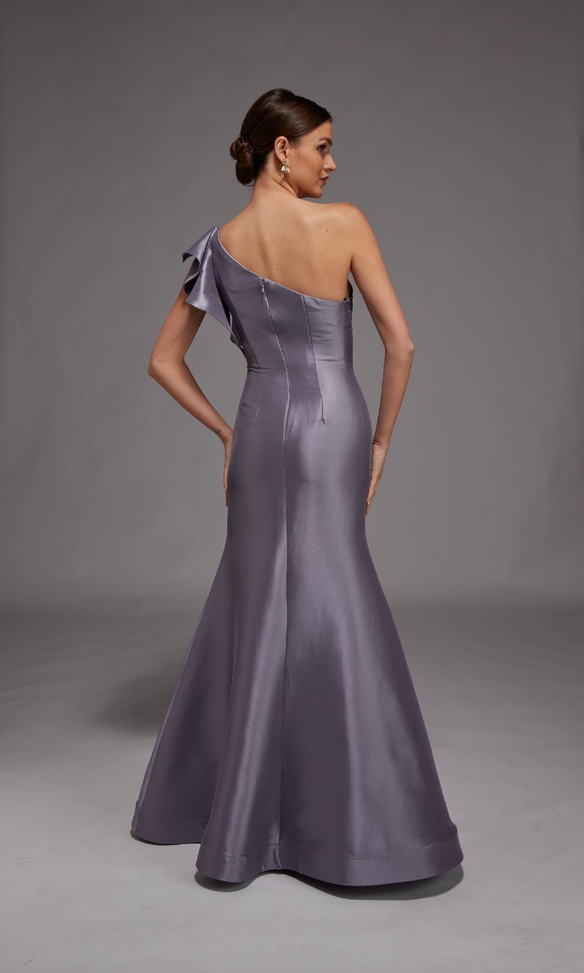 A woman stands facing away in a studio, wearing the Alyce 27826 long formal dress—a silver-lavender, one-shoulder evening gown with a fitted bodice and flared floor-length skirt.